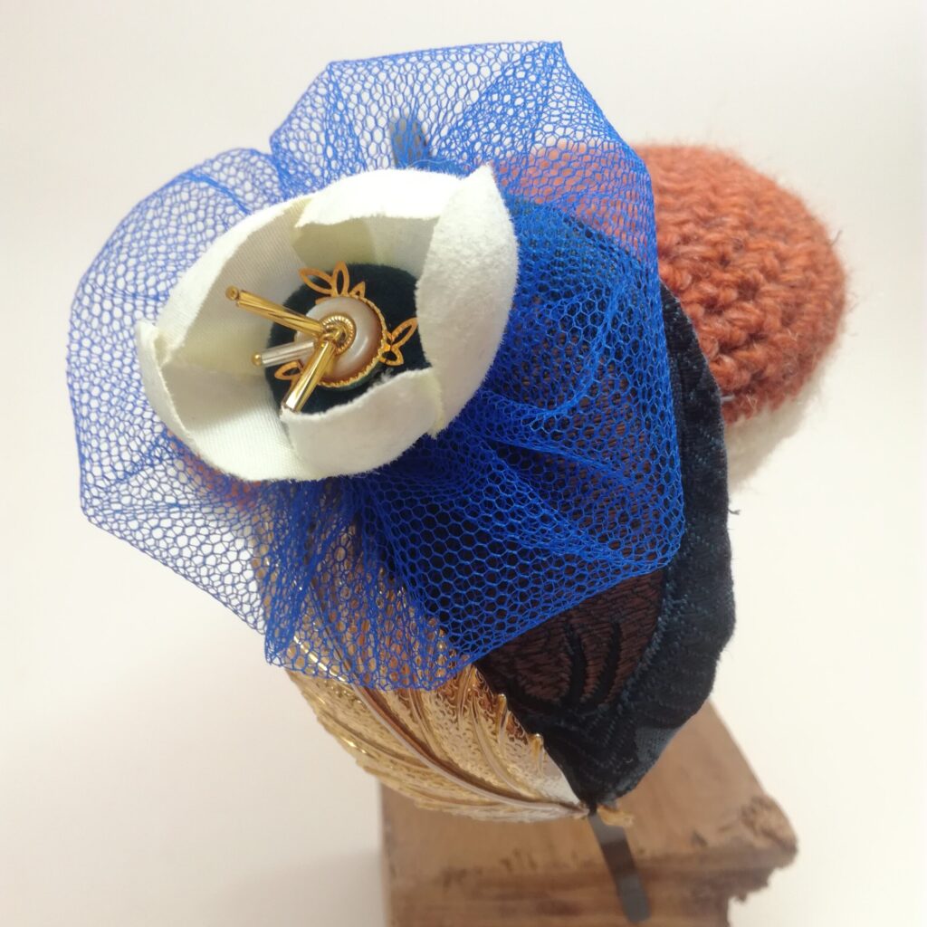 Serre-tête/bibi en métal noir composé d'un élément textile reprenant un élément d'une plume de paon  d'une fleur de résille bleu électrique à l'intérieur de laquelle se trouve une fleur articielle aux pétales blancs  une estampe métalliqe dorée filigranée et diverses perles anciennes.