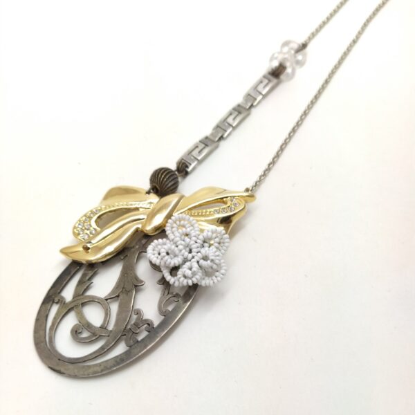 Sautoir composé de chaînes argentées composé d'un ancien monogramme argenté  d'une fleur perlée blanche et d'une broche dorée et strassée en forme de noeud.